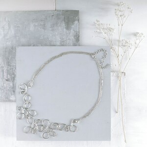 May include: A silver necklace with a delicate chain and a unique, abstract design featuring multiple interconnected loops. The necklace is displayed on a white surface with a gray background.