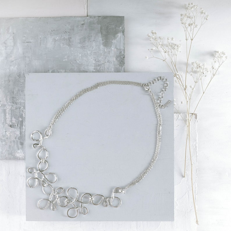 May include: A silver necklace with a delicate chain and a unique, abstract design featuring multiple interconnected loops. The necklace is displayed on a white surface with a gray background.