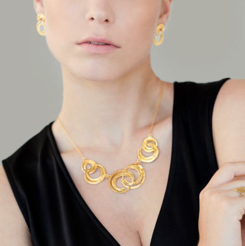 May include: A gold necklace with a design of interlocking circles. The necklace is worn by a person with a black top.