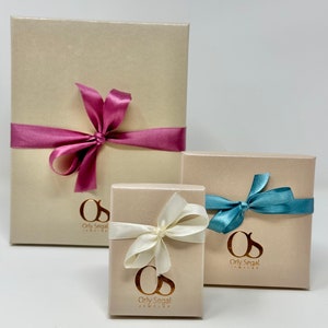 May include: Three gift boxes with beige lids and gold lettering. The largest box has a pink ribbon, the medium box has a white ribbon, and the smallest box has a blue ribbon. Each box has the text "Orly Segal Jewelry" on the front.