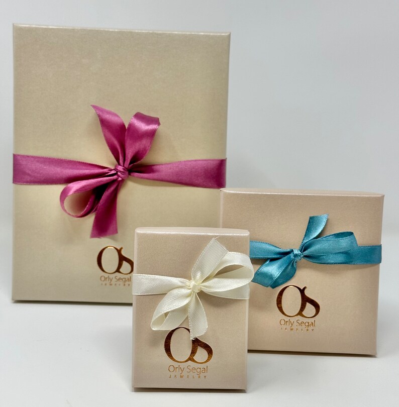 May include: Three gift boxes with beige lids and gold lettering. The largest box has a pink ribbon, the medium box has a white ribbon, and the smallest box has a blue ribbon. Each box has the text "Orly Segal Jewelry" on the front.