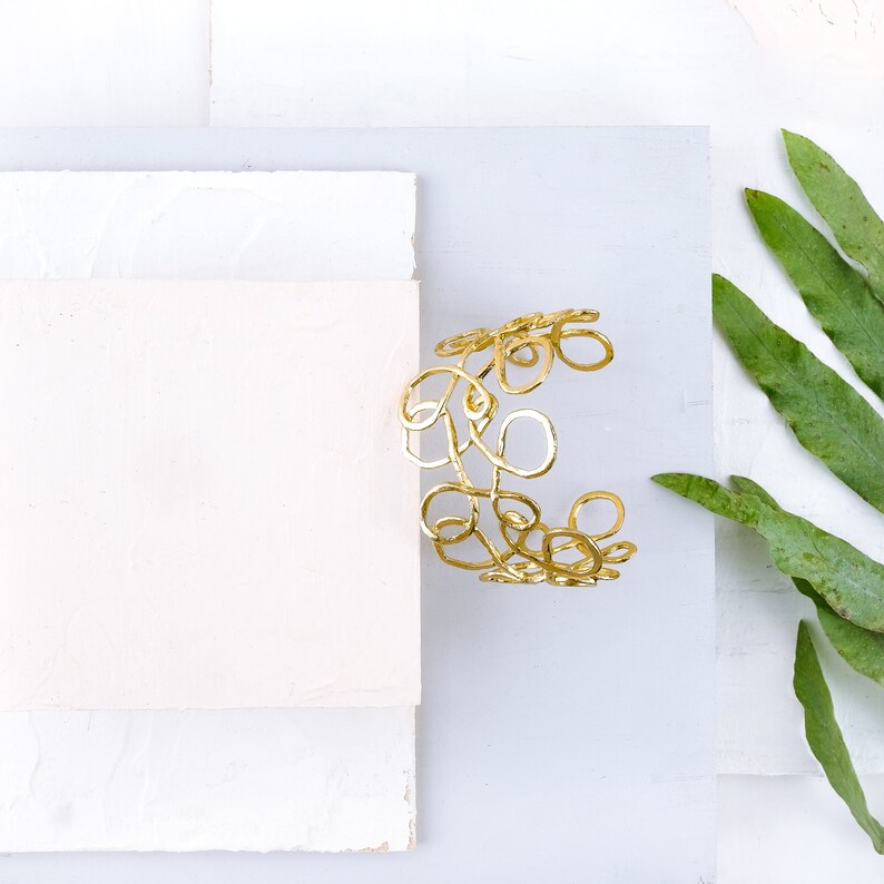 May include: A gold-toned metal bracelet with a delicate, intricate design of overlapping circles and swirls. The bracelet is on a white background.