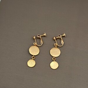 May include: A pair of gold clip-on earrings with two round discs hanging from each. The earrings are made of metal and have a shiny finish.
