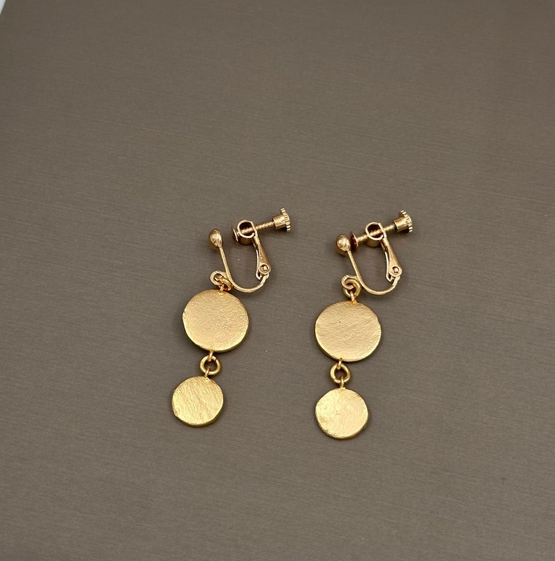 May include: A pair of gold clip-on earrings with two round discs hanging from each. The earrings are made of metal and have a shiny finish.