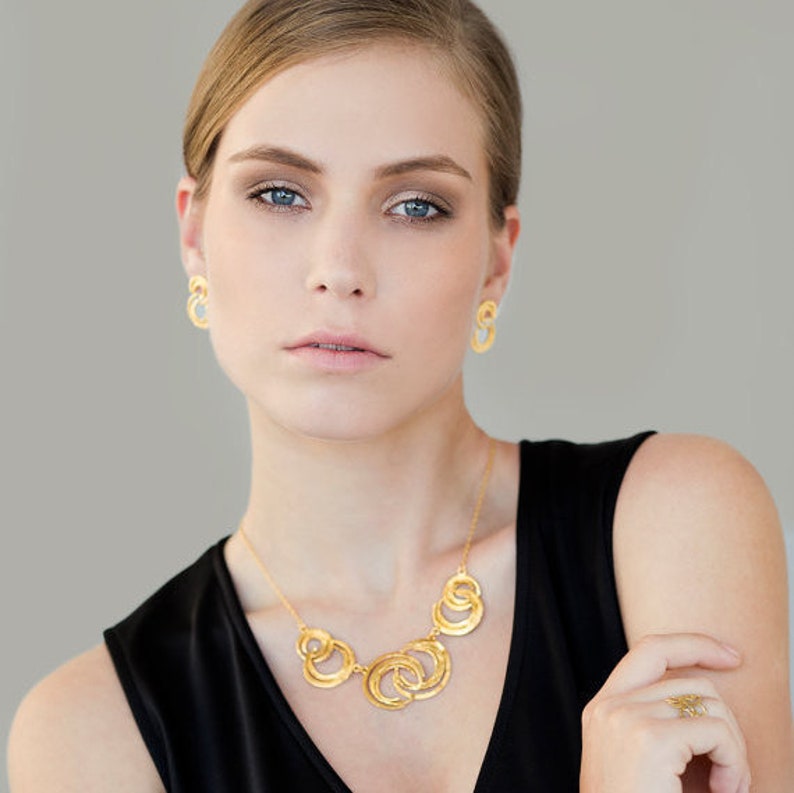 May include: A woman wearing a gold necklace and earrings with a geometric design. The necklace is made of multiple interlocking circles.