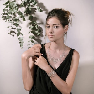 May include: A woman wearing a black sleeveless top with a silver necklace and bracelet. The necklace and bracelet are made of interconnected circles. She is also wearing silver earrings.
