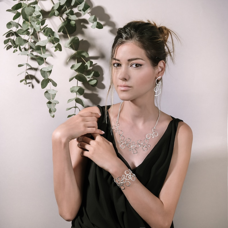 May include: A woman wearing a black sleeveless top with a silver necklace and bracelet. The necklace and bracelet are made of interconnected circles. She is also wearing silver earrings.