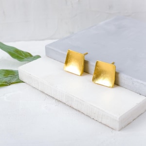 May include: A pair of gold square-shaped earrings. The earrings are made of a smooth, shiny metal and have a slightly curved shape.