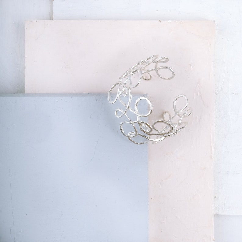 May include: A silver bracelet with a delicate, organic design. The bracelet is made of thin, twisted wire that forms a series of interconnected loops and circles.