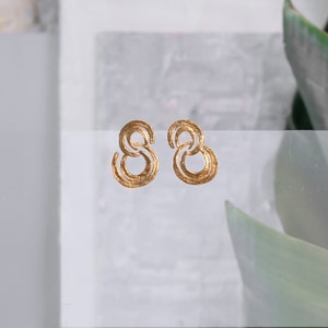 May include: A pair of gold-toned earrings with a double circle design. The earrings are textured and have a slightly hammered finish.