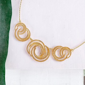 May include: A gold necklace with a geometric design featuring three interlocking circles. The circles are made of thin metal and have a textured surface.