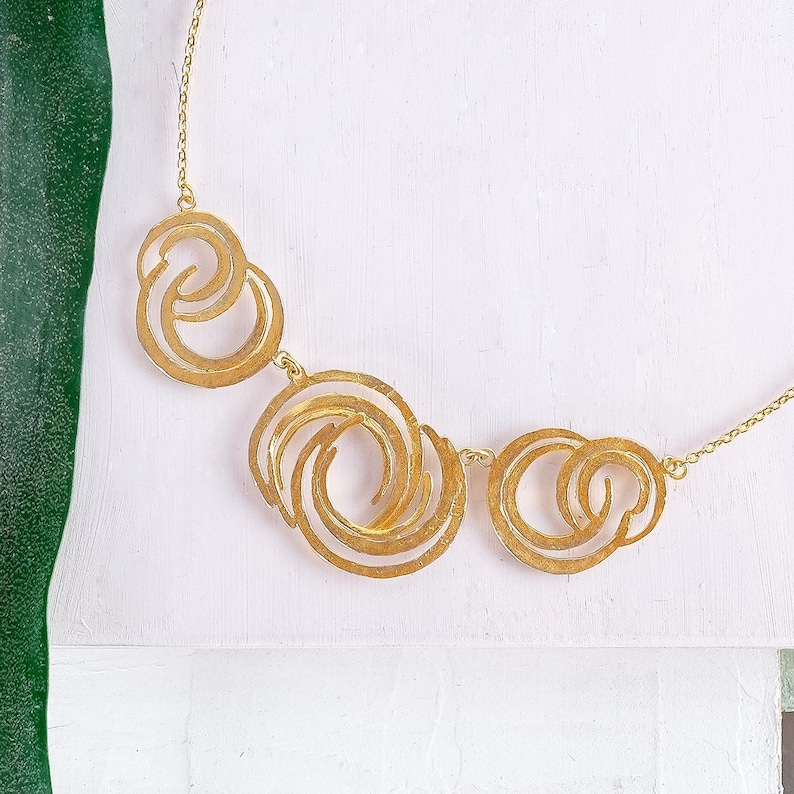 May include: A gold necklace with a geometric design featuring three interlocking circles. The circles are made of thin metal and have a textured surface.