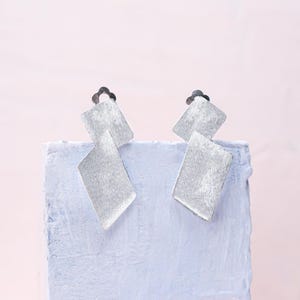 Puede incluir: Un par de pendientes de plata con un diseño geométrico. Cada pendiente presenta tres formas cuadradas superpuestas.