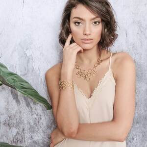 May include: A woman wearing a light beige slip dress with delicate lace trim, a gold necklace and bracelet with an intricate design. She is posing with her hand on her chin, looking at the camera.