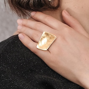 Könnte beinhalten: Ein rechteckiger Ring in Goldton mit strukturierter Oberfläche. Der Ring wird an einem Finger getragen.