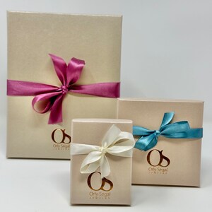 May include: Three gift boxes with beige lids and gold lettering. The largest box has a pink ribbon, the medium box has a white ribbon, and the smallest box has a blue ribbon. Each box has the text "Orly Segal Jewelry" on the front.