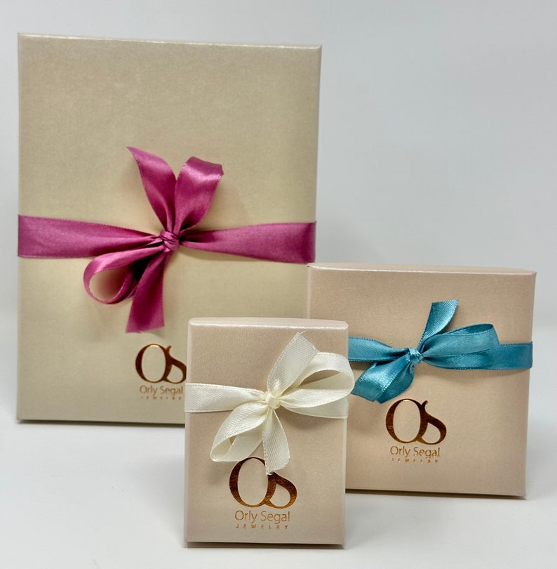 May include: Three gift boxes with beige lids and gold lettering. The largest box has a pink ribbon, the medium box has a white ribbon, and the smallest box has a blue ribbon. Each box has the text "Orly Segal Jewelry" on the front.