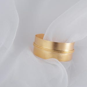 May include: A gold-colored metal bangle bracelet with a smooth, brushed finish. The bracelet is thick and wide, with a subtle groove around the center.