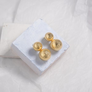 Op de afbeelding: Een paar gouden oorbellen met twee ronde, getextureerde bollen aan elke oorbel.