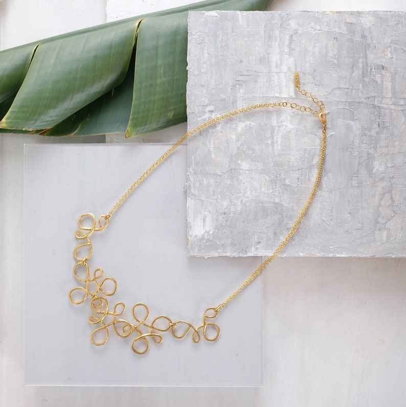 May include: A gold necklace with a delicate, intricate design. The necklace is made of thin, twisted wire that forms a series of loops and swirls. The necklace is displayed on a white surface with a green leaf in the background.