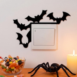 May include: A black decorative wall decal featuring three bat silhouettes positioned around a square white light switch. The bats are arranged with two flying above the switch and one hanging vertically along the left side of the switch plate.