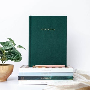May include: A stack of green, blue, and white notebooks with a pink pen on top. The notebooks have the word "NOTEBOOK" printed on the front. A green notebook with the word "NOTEBOOK" printed on the front is behind the stack.