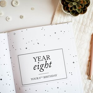 May include: A white journal with a black star pattern and the text "YEAR eight YOUR 8TH BIRTHDAY" on the cover. A pink pen is lying on a white surface next to the journal.