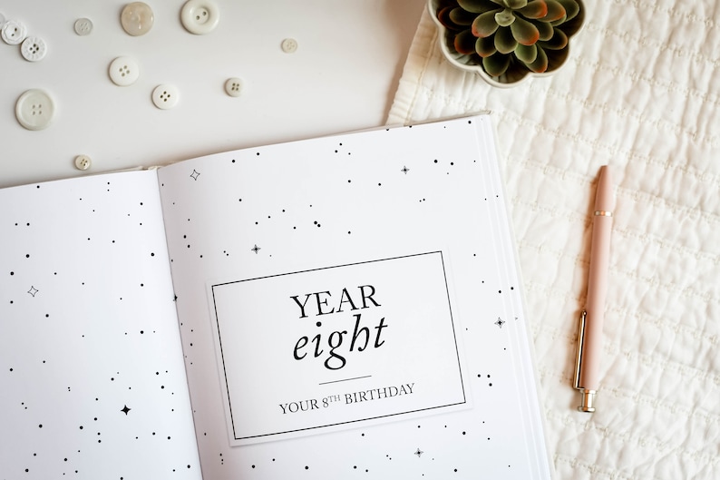 May include: A white journal with a black star pattern and the text "YEAR eight YOUR 8TH BIRTHDAY" on the cover. A pink pen is lying on a white surface next to the journal.