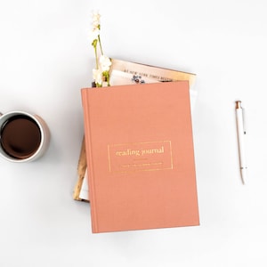 K&ouml;nnte beinhalten: Ein rosafarbenes Lesetagebuch mit goldener Aufschrift "reading journal" auf dem Einband. Das Tagebuch liegt auf einem Stapel B&uuml;cher und ein wei&szlig;er Stift liegt auf einer wei&szlig;en Oberfl&auml;che.