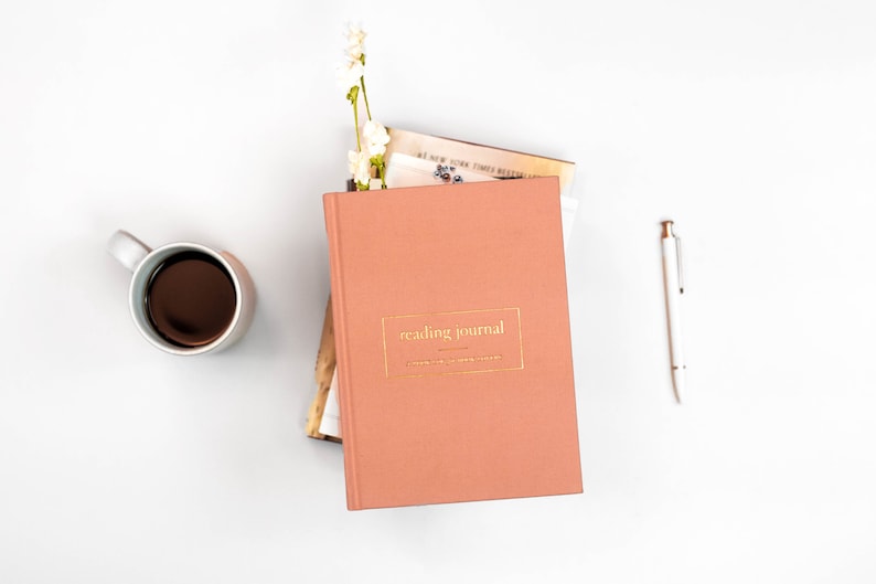K&ouml;nnte beinhalten: Ein rosafarbenes Lesetagebuch mit goldener Aufschrift "reading journal" auf dem Einband. Das Tagebuch liegt auf einem Stapel B&uuml;cher und ein wei&szlig;er Stift liegt auf einer wei&szlig;en Oberfl&auml;che.