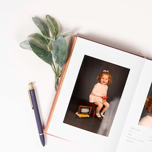 K&ouml;nnte beinhalten: Ein offenes Fotoalbum mit einem rosa Einband. Die linke Seite zeigt ein Foto eines jungen M&auml;dchens in einem rosa Kleid, das auf einem kleinen roten Hocker sitzt. Die rechte Seite zeigt ein Foto eines jungen M&auml;dchens mit roten Haaren. Das Fotoalbum hat einen wei&szlig;en Hintergrund und einen wei&szlig;en Rahmen um jedes Foto.