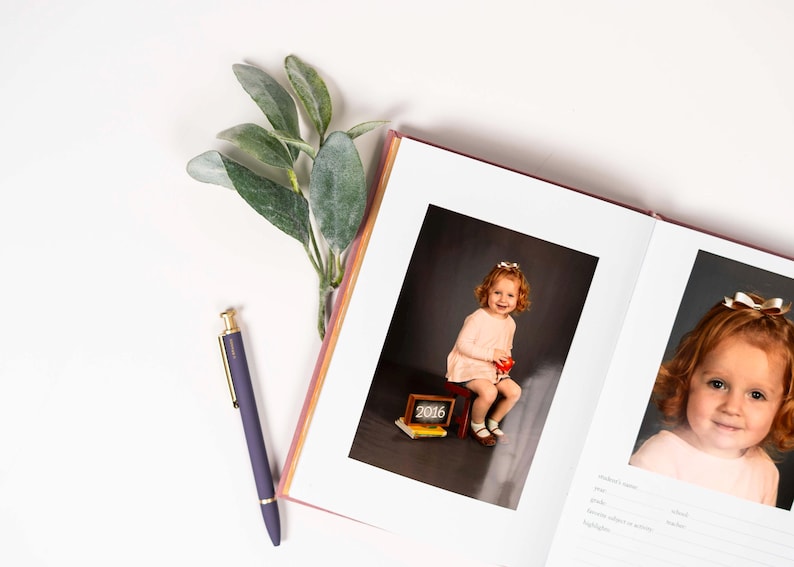 K&ouml;nnte beinhalten: Ein offenes Fotoalbum mit einem rosa Einband. Die linke Seite zeigt ein Foto eines jungen M&auml;dchens in einem rosa Kleid, das auf einem kleinen roten Hocker sitzt. Die rechte Seite zeigt ein Foto eines jungen M&auml;dchens mit roten Haaren. Das Fotoalbum hat einen wei&szlig;en Hintergrund und einen wei&szlig;en Rahmen um jedes Foto.