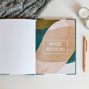 K&ouml;nnte beinhalten: Ein offenes Buch mit wei&szlig;em Hintergrund und dem Text "BOOK REVIEWS" auf dem Einband. Das Buch liegt auf einer wei&szlig;en Oberfl&auml;che mit einer grauen Strickdecke, zwei wei&szlig;en Kerzen und einem rosa Stift.