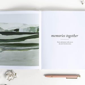 May include: Open book with a landscape painting on the left page and text that reads "memories together" on the right. A pink pen rests below the right page. White flowers are scattered around the book.