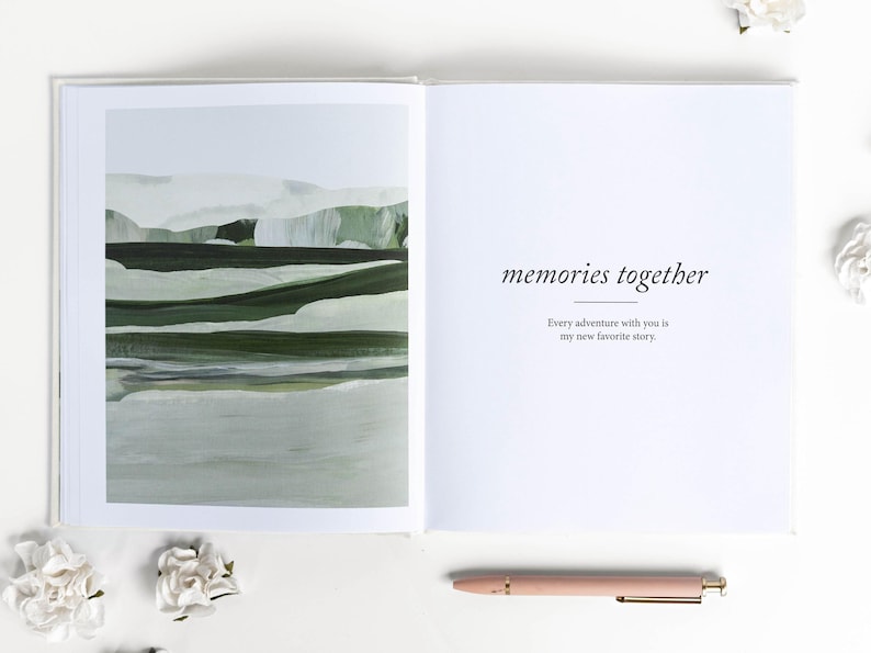 May include: Open book with a landscape painting on the left page and text that reads "memories together" on the right. A pink pen rests below the right page. White flowers are scattered around the book.