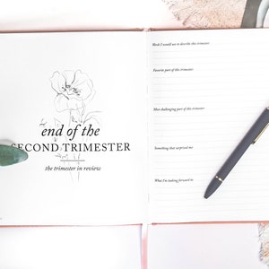 May include: A white journal with a black floral illustration and the text "end of the SECOND TRIMESTER the trimester in review". The journal has lined pages for writing and a black pen is lying on the open pages.