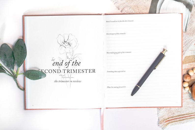 May include: A white journal with a black floral illustration and the text "end of the SECOND TRIMESTER the trimester in review". The journal has lined pages for writing and a black pen is lying on the open pages.