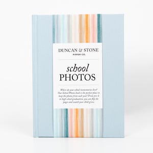 K&ouml;nnte beinhalten: Ein hellblaues Fotoalbum mit dem Titel "school photos" und einem bunten gestreiften Design auf dem Cover. Der Text auf dem Cover lautet "Wo leben deine Schulerinnerungen? Unser Schulfotoalbum ist der perfekte Ort, um die Fotos aus jedem Jahr aufzubewahren! Von der Vorschule bis zum Abitur kannst du die Seiten umbl&auml;ttern und zusehen, wie dein Kind w&auml;chst."