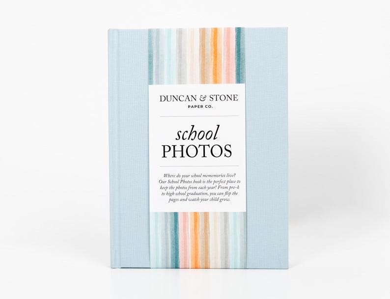 K&ouml;nnte beinhalten: Ein hellblaues Fotoalbum mit dem Titel "school photos" und einem bunten gestreiften Design auf dem Cover. Der Text auf dem Cover lautet "Wo leben deine Schulerinnerungen? Unser Schulfotoalbum ist der perfekte Ort, um die Fotos aus jedem Jahr aufzubewahren! Von der Vorschule bis zum Abitur kannst du die Seiten umbl&auml;ttern und zusehen, wie dein Kind w&auml;chst."