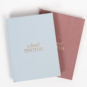 K&ouml;nnte beinhalten: Zwei Fotoalben, eines hellblau und eines staubig rosa, mit dem Text "school photos" in goldenen Buchstaben auf der Vorderseite.