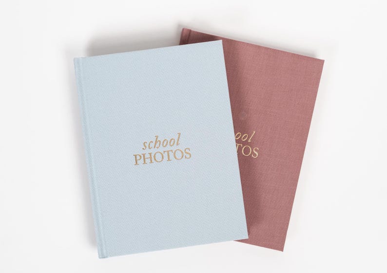 K&ouml;nnte beinhalten: Zwei Fotoalben, eines hellblau und eines staubig rosa, mit dem Text "school photos" in goldenen Buchstaben auf der Vorderseite.