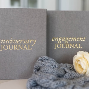 May include: Two gray linen-covered journals with gold lettering. The journal on the left says "anniversary JOURNAL" and the journal on the right says "engagement JOURNAL".