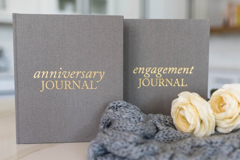 May include: Two gray linen-covered journals with gold lettering. The journal on the left says "anniversary JOURNAL" and the journal on the right says "engagement JOURNAL".