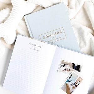 May include: A light blue journal titled "A Dog's Life" with gold lettering, open to a page with the text "Gotcha Story." Two dog photos are on the open page. White dog bone toys are in the background.