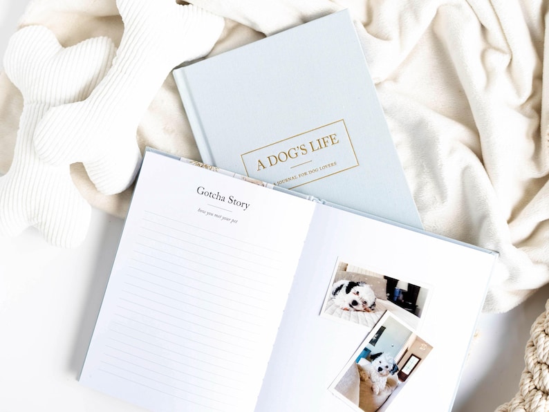 May include: A light blue journal titled "A Dog's Life" with gold lettering, open to a page with the text "Gotcha Story." Two dog photos are on the open page. White dog bone toys are in the background.