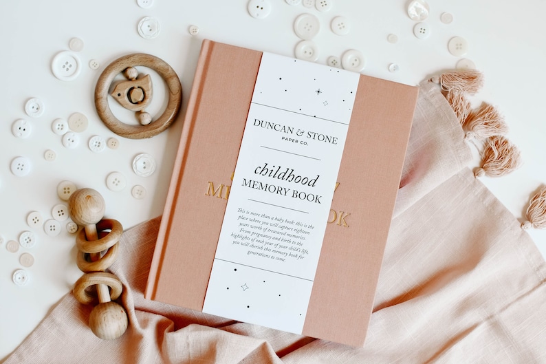 May include: A pink book with the title "childhood memory book" and the text "This is more than a baby book, this is the place where you will capture eighteen years worth of treasured memories. From pregnancy and birth to the highlights of each year of your child's life, you will cherish this memory book for generations to come."