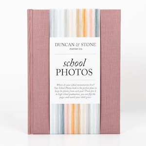 K&ouml;nnte beinhalten: Ein rosa Fotoalbum mit einem gestreiften Einband und dem Text "Duncan & Stone Paper Co. school PHOTOS" auf der Vorderseite. Der Text unter dem Titel lautet "Wo leben deine Schulerinnerungen? Unser Schulfotoalbum ist der perfekte Ort, um die Fotos aus jedem Jahr aufzubewahren! Von der Vorschule bis zum Abitur kannst du die Seiten umbl&auml;ttern und zusehen, wie dein Kind w&auml;chst."