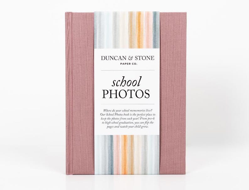 K&ouml;nnte beinhalten: Ein rosa Fotoalbum mit einem gestreiften Einband und dem Text "Duncan & Stone Paper Co. school PHOTOS" auf der Vorderseite. Der Text unter dem Titel lautet "Wo leben deine Schulerinnerungen? Unser Schulfotoalbum ist der perfekte Ort, um die Fotos aus jedem Jahr aufzubewahren! Von der Vorschule bis zum Abitur kannst du die Seiten umbl&auml;ttern und zusehen, wie dein Kind w&auml;chst."