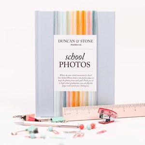 May include: A light blue photo album with the text "school PHOTOS" on the cover. The album is bound with a colorful striped ribbon. A ruler, stapler, and various stationery items are scattered in front of the album.
