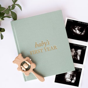 May include: A green baby memory book with gold lettering that reads "baby's first year". A wooden rattle and ultrasound photos are also in the image.